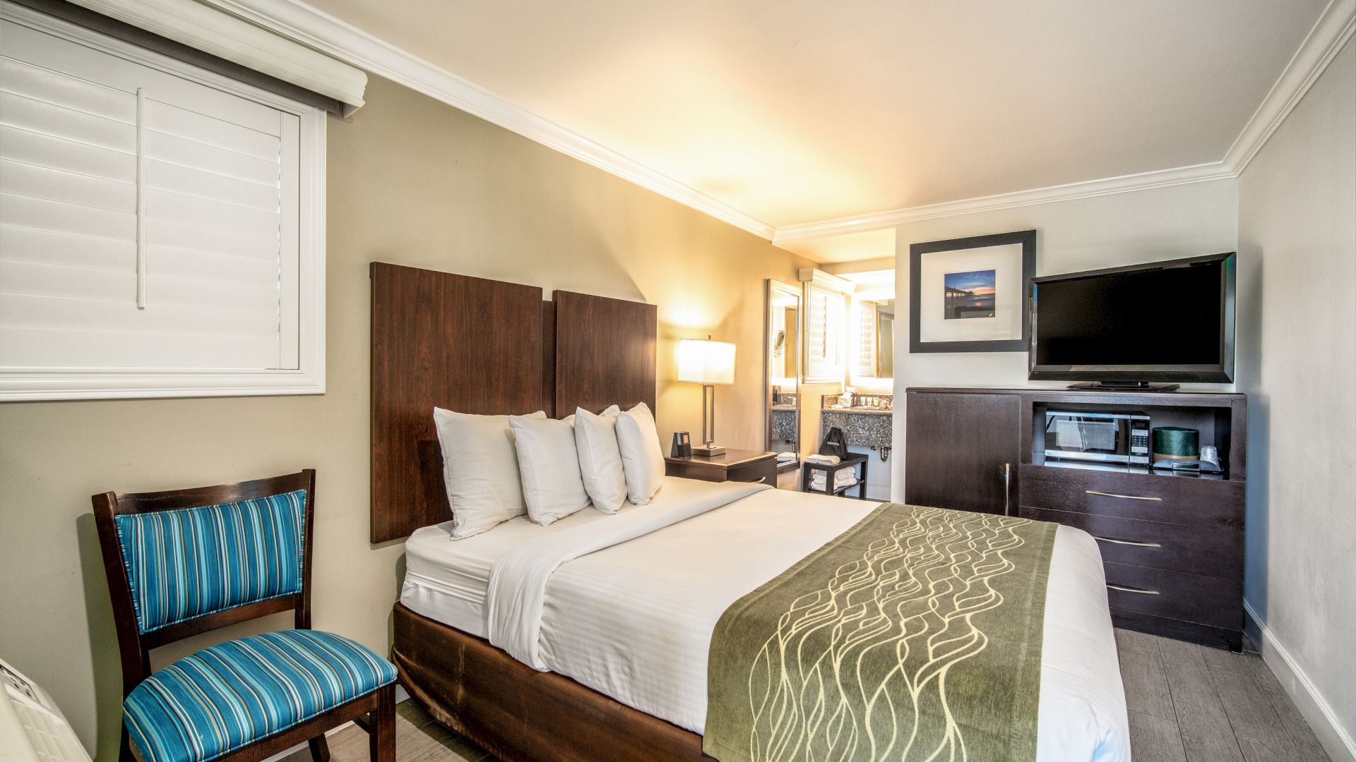A neatly arranged hotel room with a double bed, a blue-striped chair, a TV atop a dresser, and a window with shutters ending the sentence.