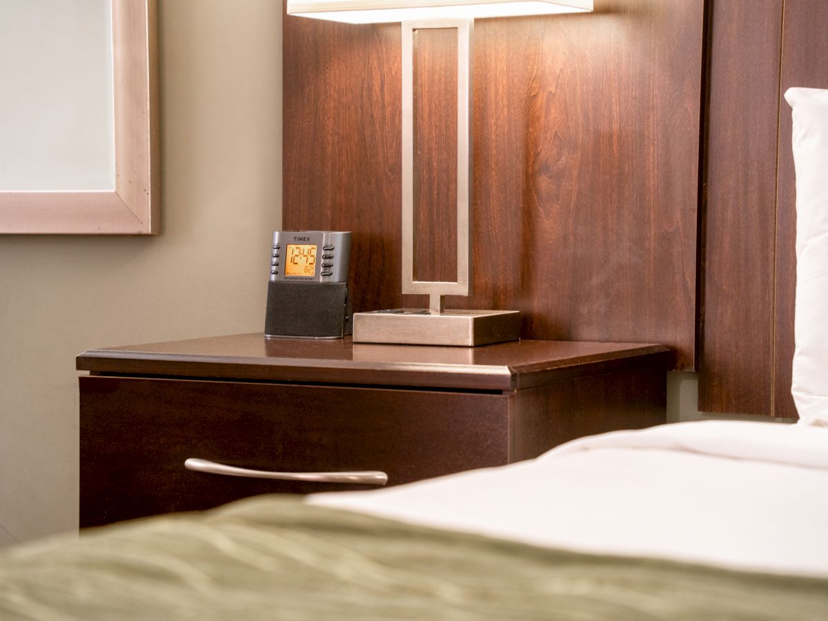 A bedside table with a lamp, an alarm clock, and part of a bed are in the image.