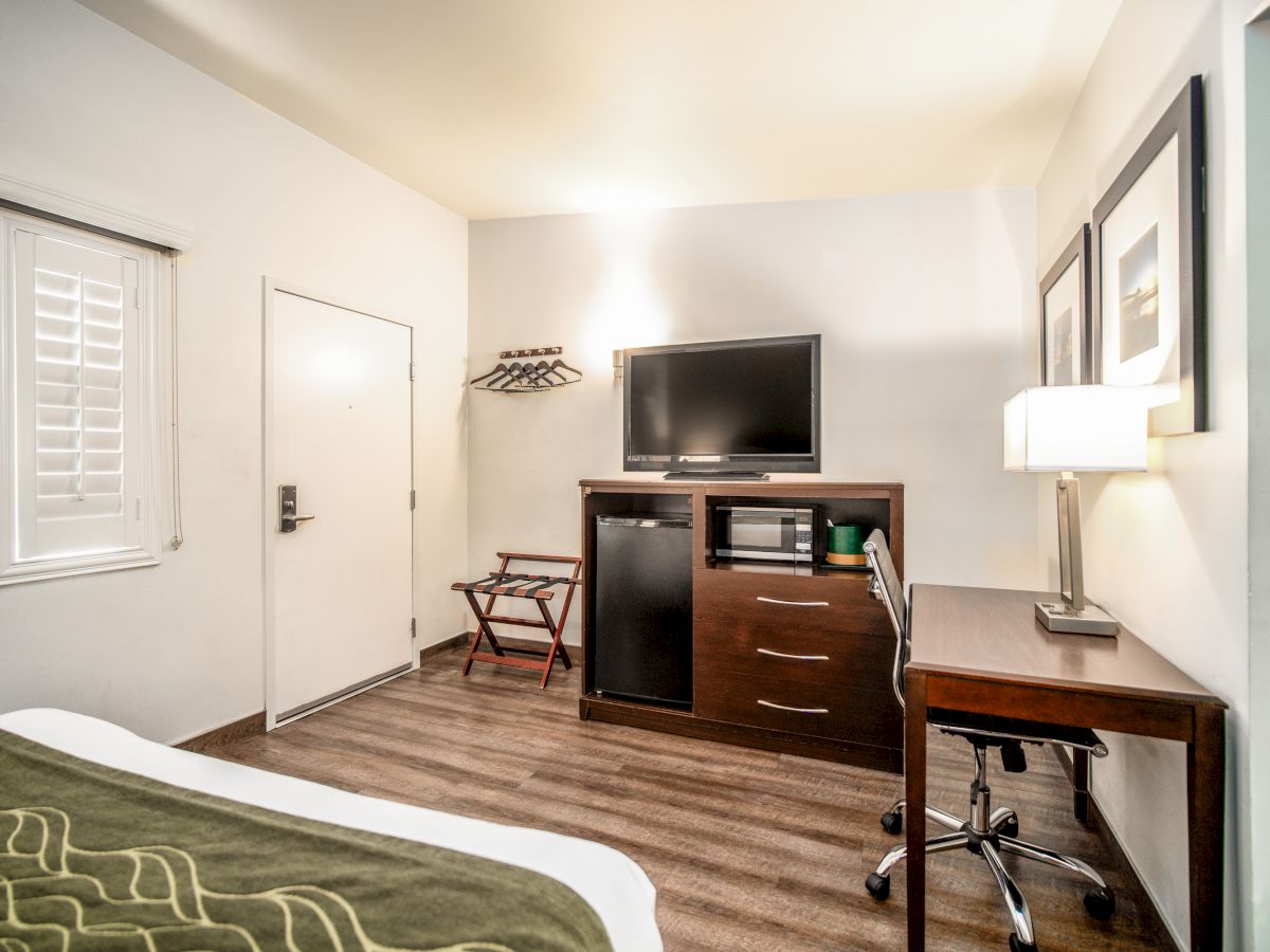 A hotel room with a bed, TV, desk, lamp, and storage unit containing a microwave and fridge, decorated with framed pictures and an entry door.