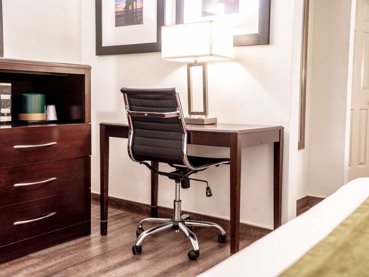 A room with a wooden desk, black office chair, desk lamp, framed pictures on the wall, a dresser, and a door. The floor is hardwood.