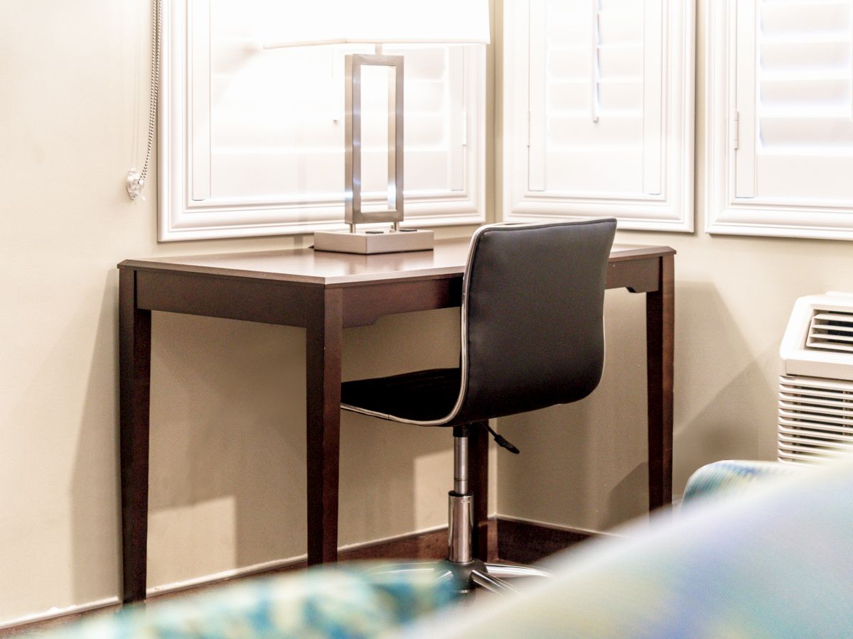 A small wooden desk with a modern lamp and a black chair is placed against a wall with shuttered windows, beside an air conditioning unit.