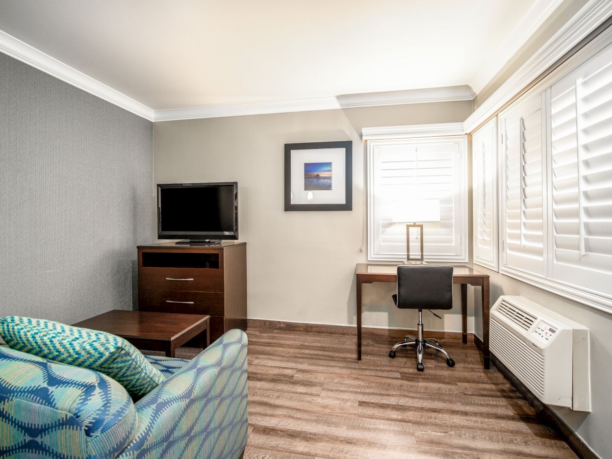 A modern room with a TV on a cabinet, a desk with a chair, an armchair, a lamp, wall artwork, window shutters, and a hardwood floor.