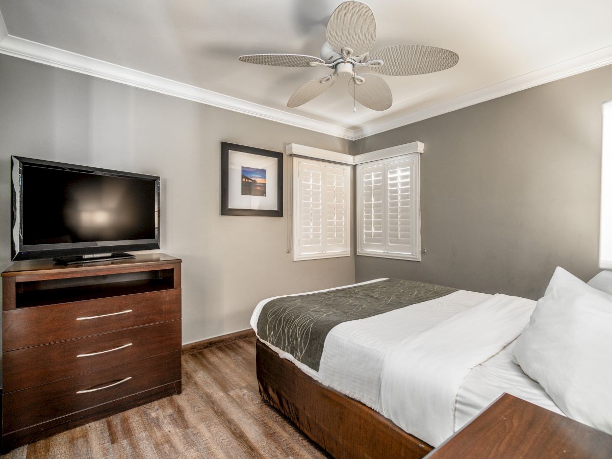 The image shows a bedroom with a bed, a TV on a dresser, a ceiling fan, a framed picture, and windows with white shutters.