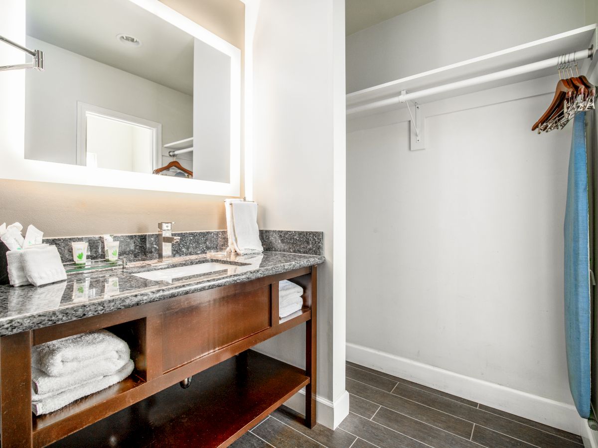 A bathroom with a well-lit vanity, a large mirror, countertop items, and an adjacent closet with hangers and a blue ironing board is shown.