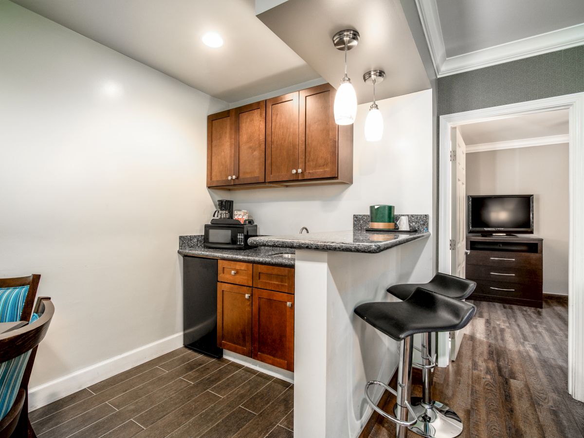 A small kitchen area with wooden cabinets, a microwave, and bar stools next to a dining space; a doorway shows a TV in another room.