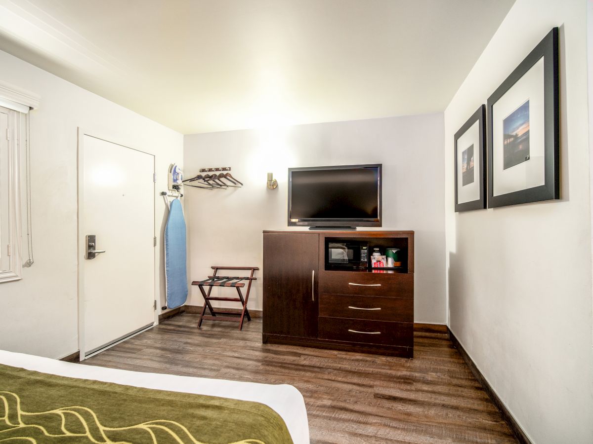 The image shows a hotel room with a TV on a dresser, microwave, two framed pictures on the wall, an ironing board, and a luggage rack.