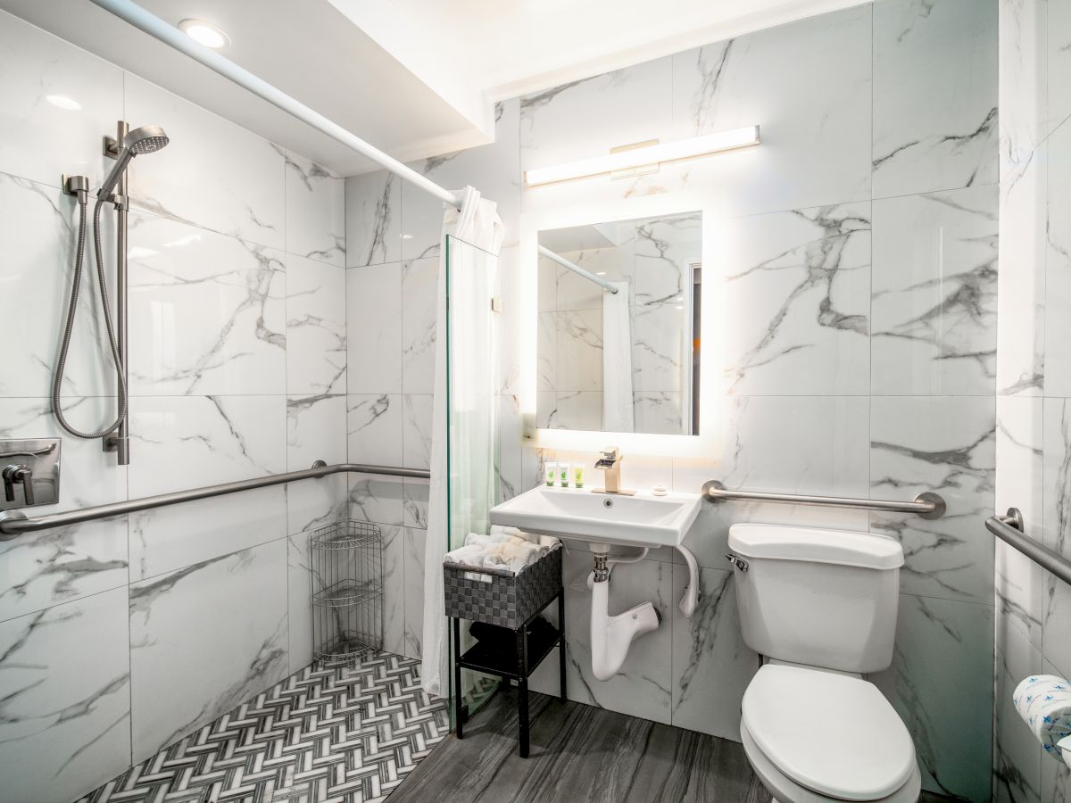 This image shows a modern bathroom with a walk-in shower, toilet, sink with a vanity mirror, and grab bars along the walls, all in a marble design.