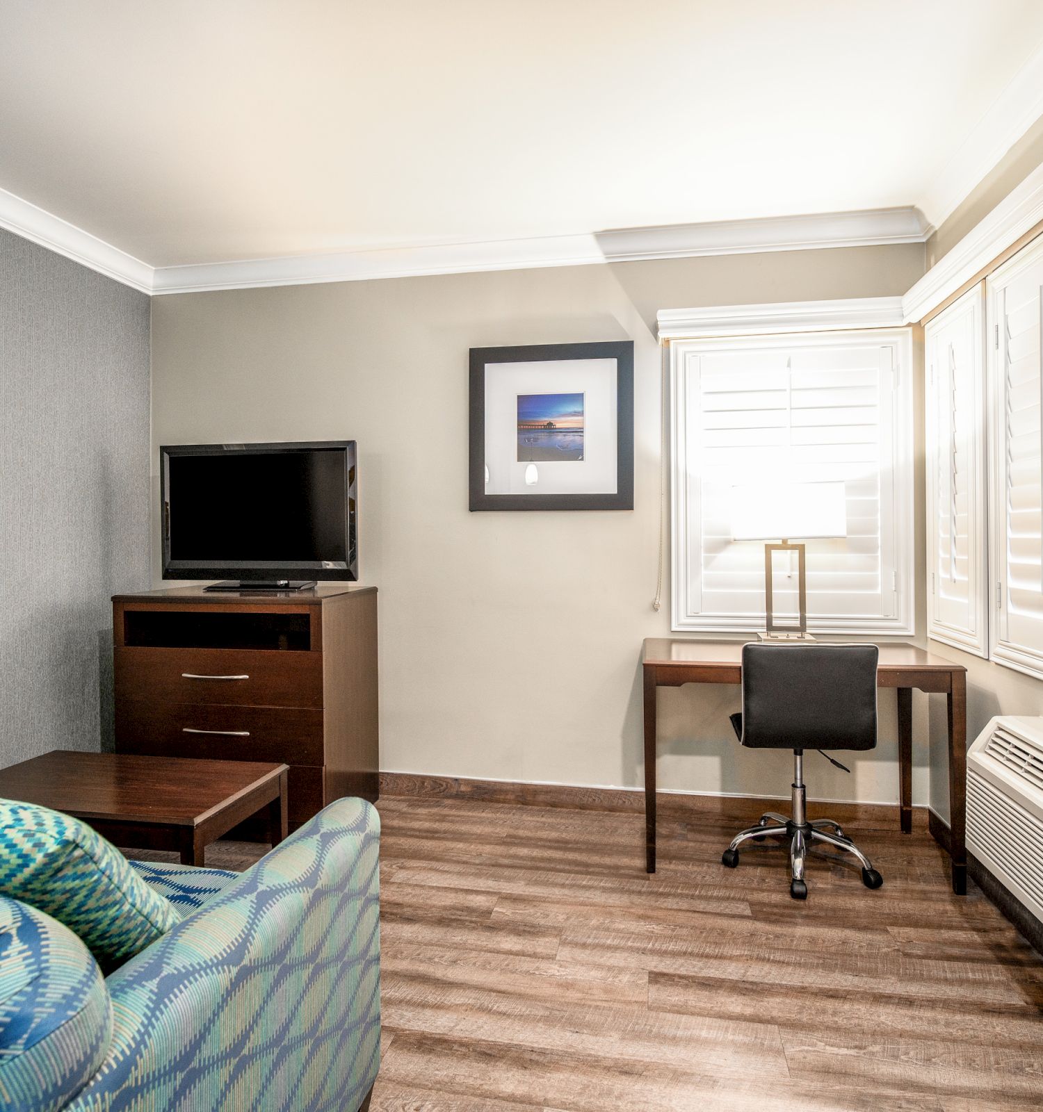 A room with a TV on a dresser, a desk with a lamp and chair, a couch with patterned pillows, and a window with shutters, all on wooden flooring.