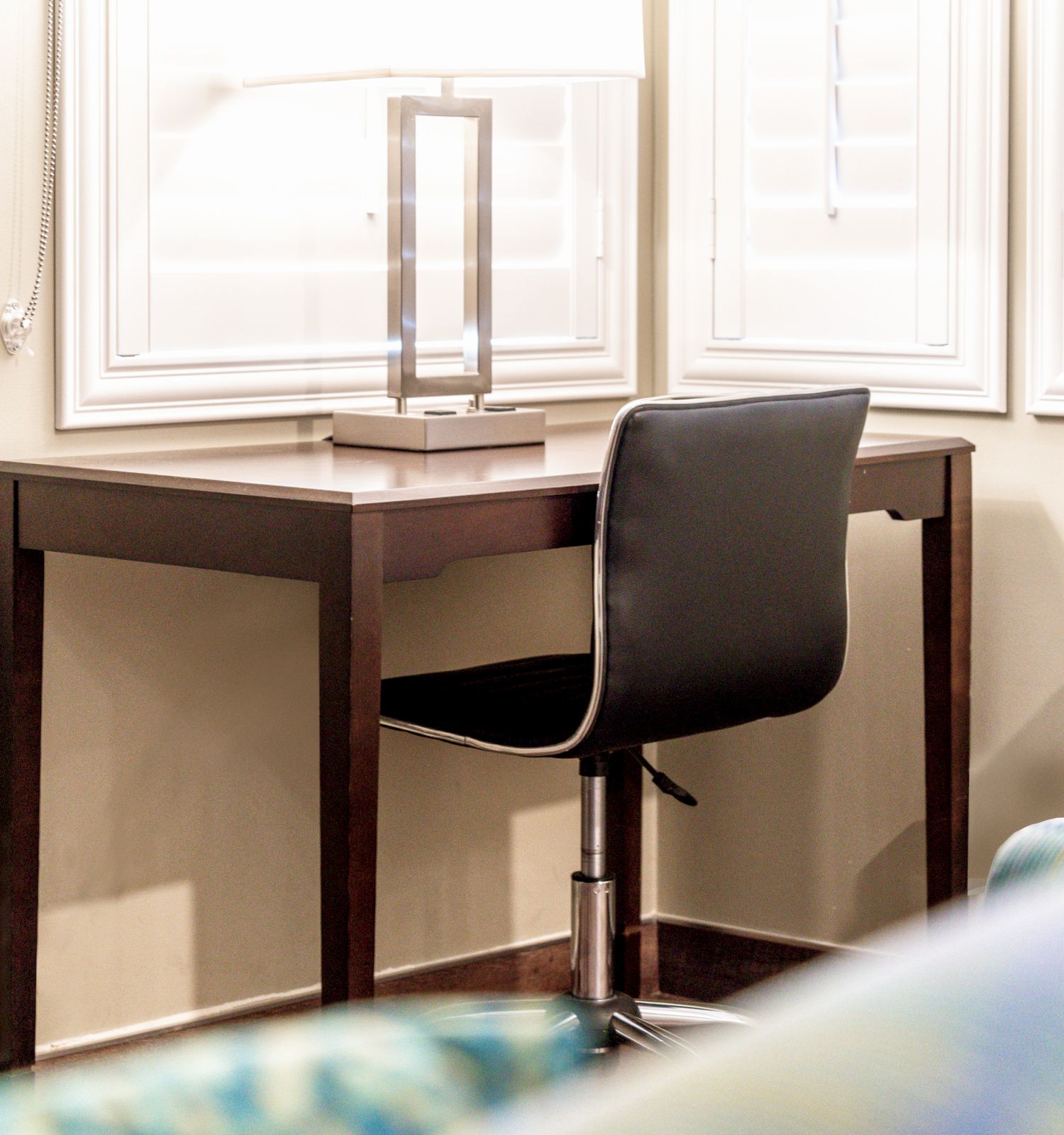 The image shows a small wooden desk with a modern lamp and a black chair, situated in front of two windows with closed blinds.