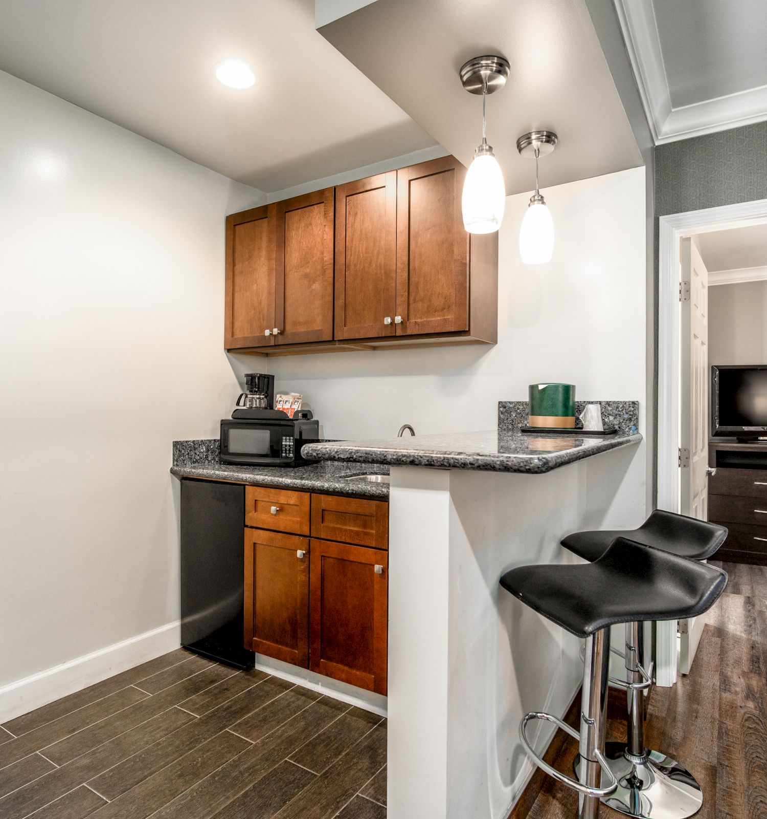 A kitchenette with wooden cabinets, barstools, and a dining table. There's also a view into an adjacent room with a TV and dresser.