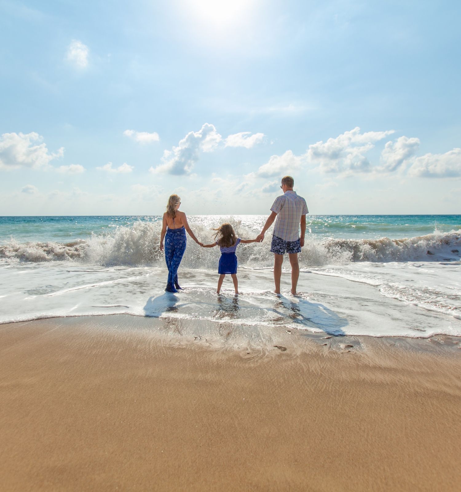 A family of three standing hand in hand at the edge of the ocean on a sunny beach, with waves gently crashing against the shore.