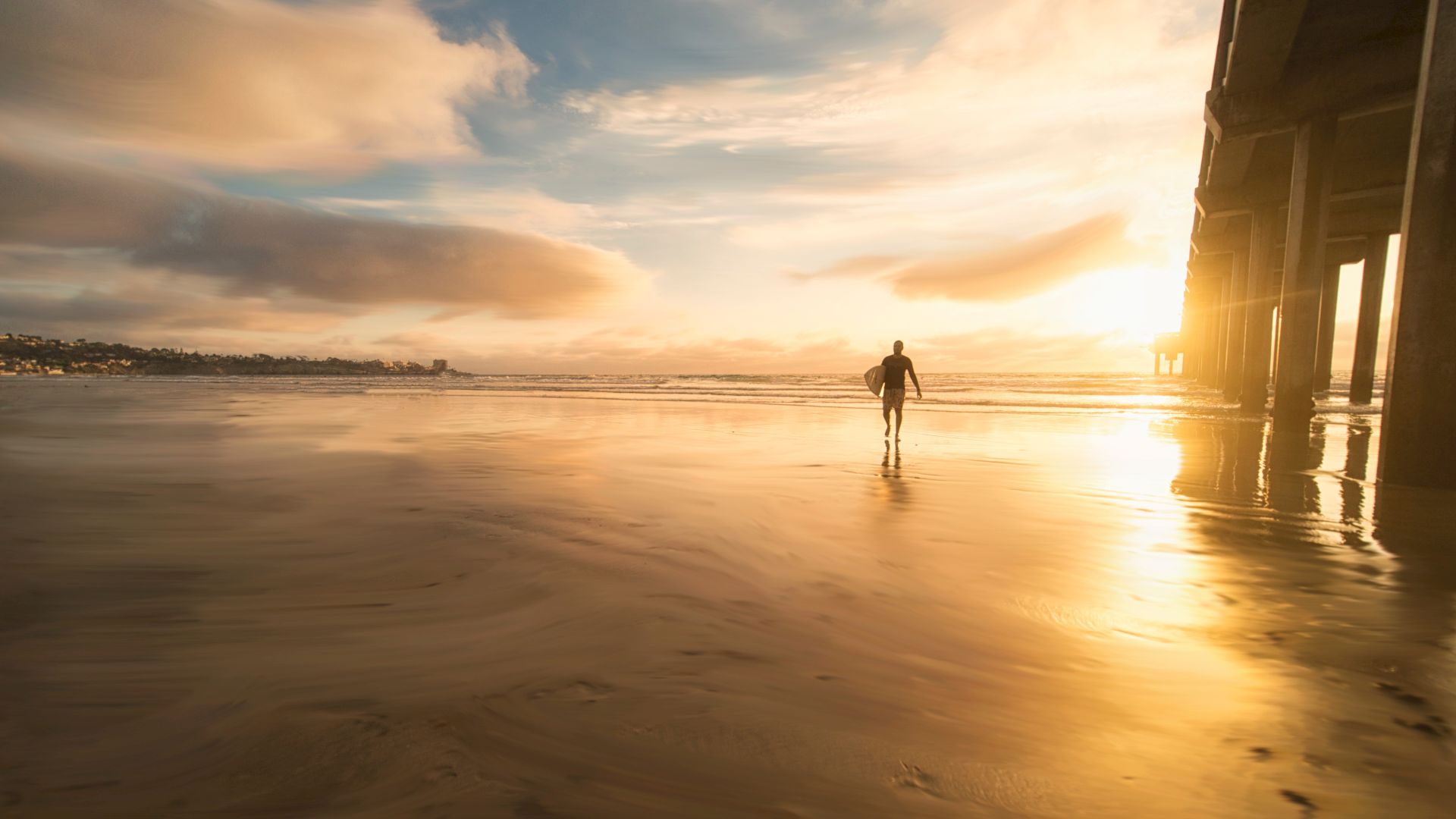 A solitary figure walks on a beach during sunset with a pier to the right and dramatic clouds in the sky's background.