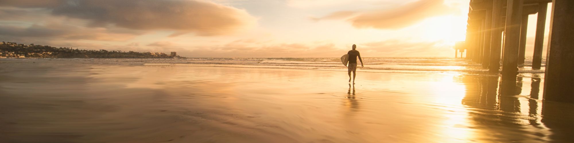 A solitary figure walks on a beach during sunset with a pier to the right and dramatic clouds in the sky's background.