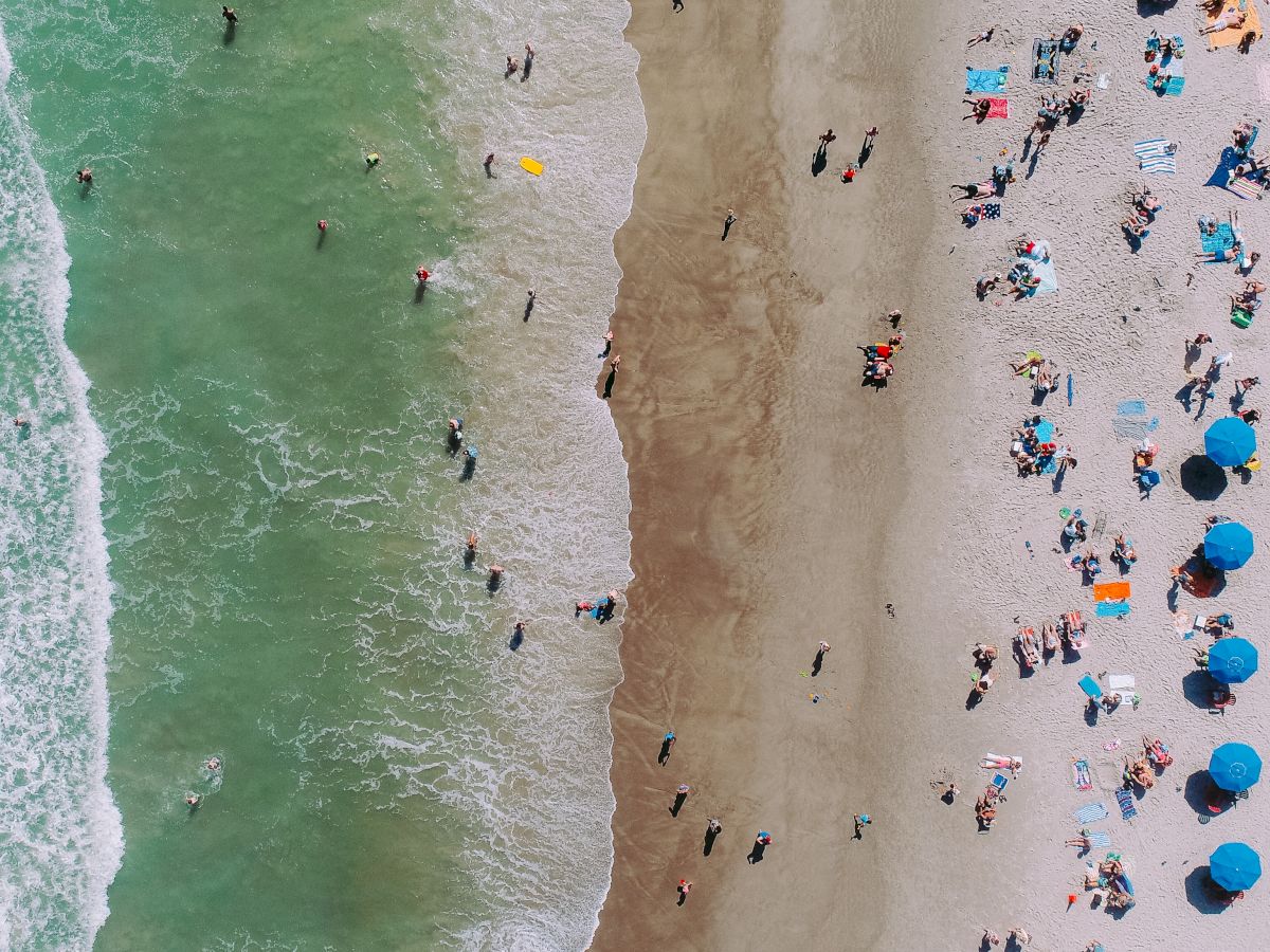 An aerial view of a beach with people swimming in the ocean and sunbathing under umbrellas. The image captures waves hitting the shore.