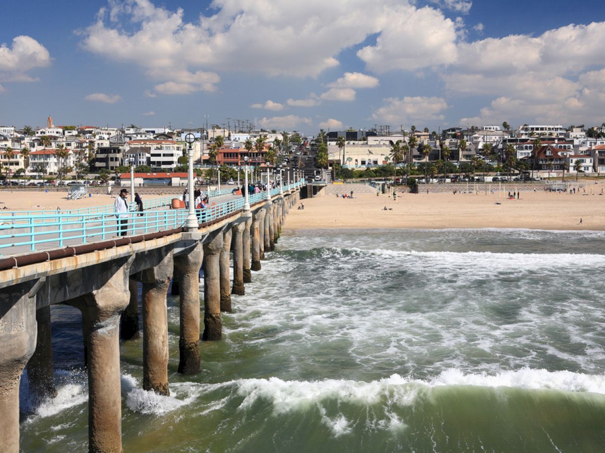 A coastal beach scene with a long pier extending into the ocean, waves crashing against it, and a sandy beach with buildings and people in the background.