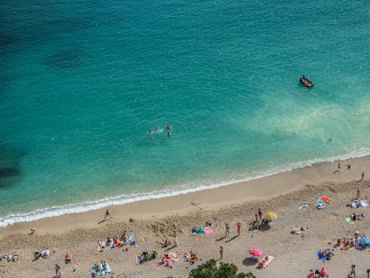 An aerial view of a sandy beach with people sunbathing, umbrellas dotted around, and a few swimmers in clear turquoise water.