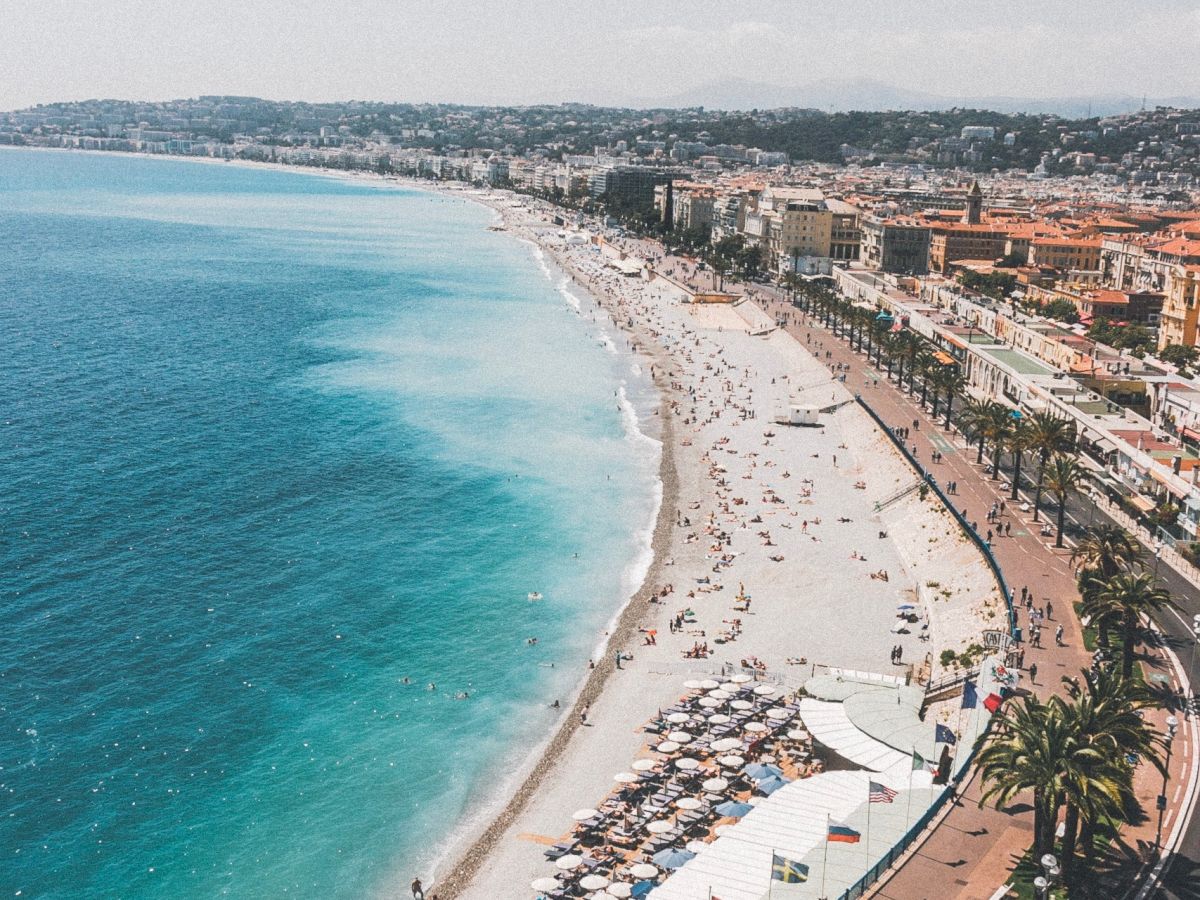 A coastal cityscape with a sunny beach lined with umbrellas and buildings. A road runs along the shoreline with palm trees and ocean views.