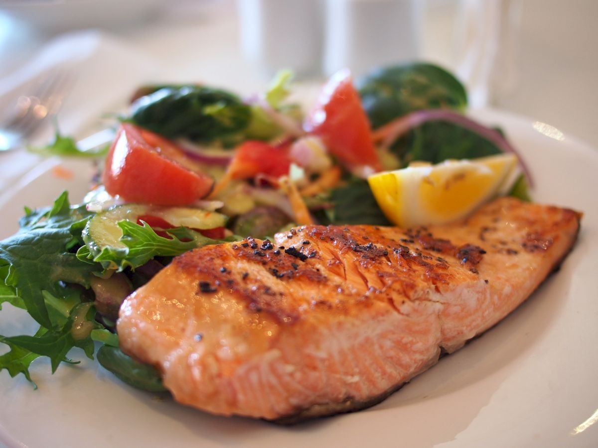 The image shows a grilled salmon fillet served with a fresh mixed salad including tomatoes, red onions, and a lemon wedge on a white plate.