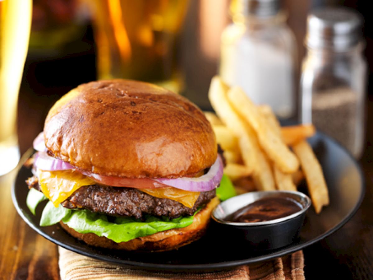 The image shows a plate with a cheeseburger, lettuce, tomato, and onion, accompanied by fries and a small container of sauce, along with a beer in the background.