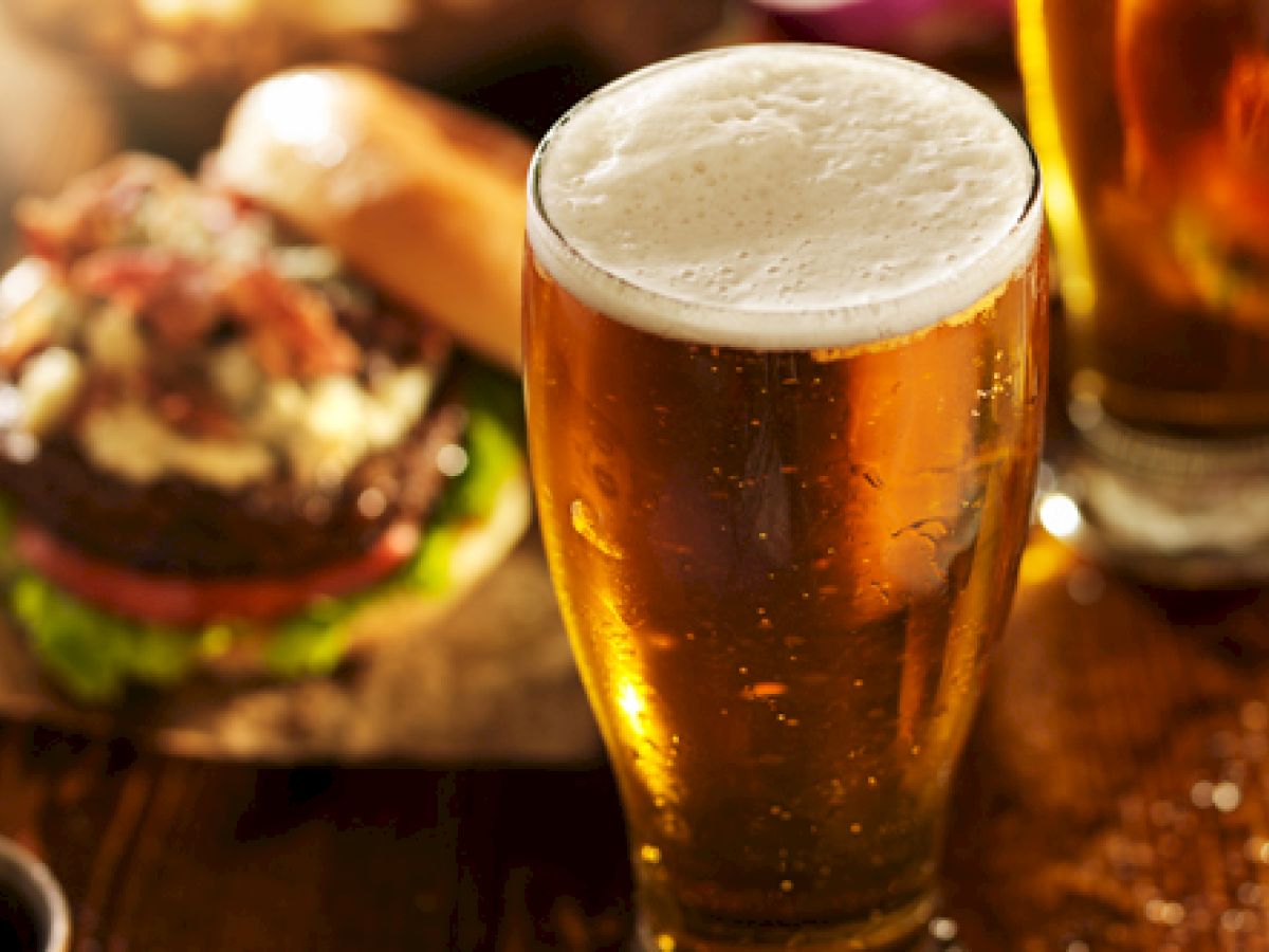 The image shows a glass of beer and a juicy burger with various toppings on a wooden table. Another glass of beer is in the background.