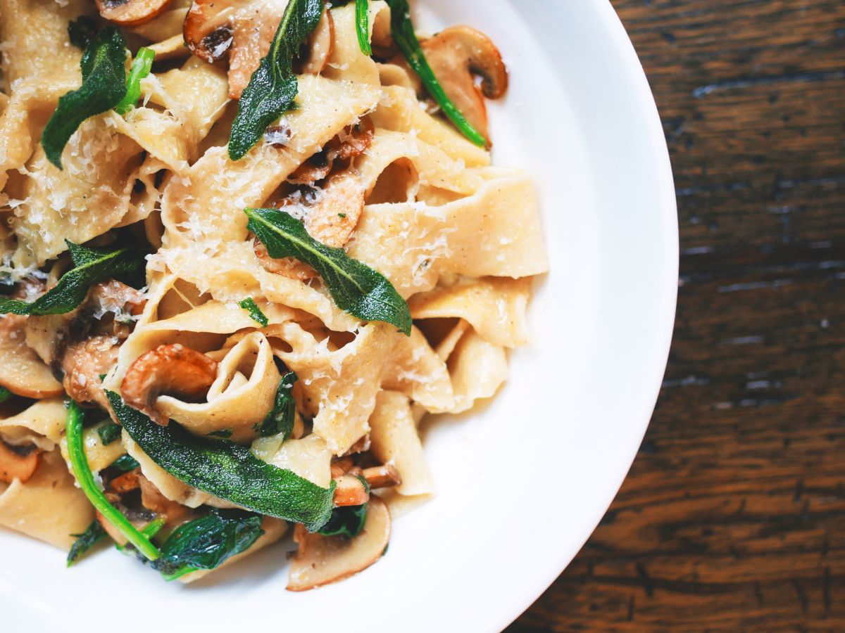 A white dish filled with wide pasta noodles, mushrooms, and leafy greens is shown, placed on a wooden table surface.