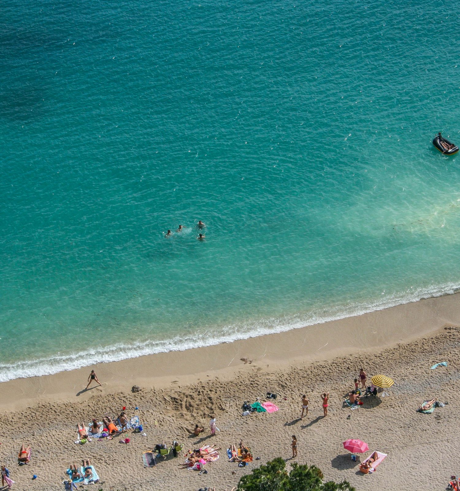 An aerial view of a beach with people sunbathing on the sand and swimming in the clear blue water, along with a small boat further out.