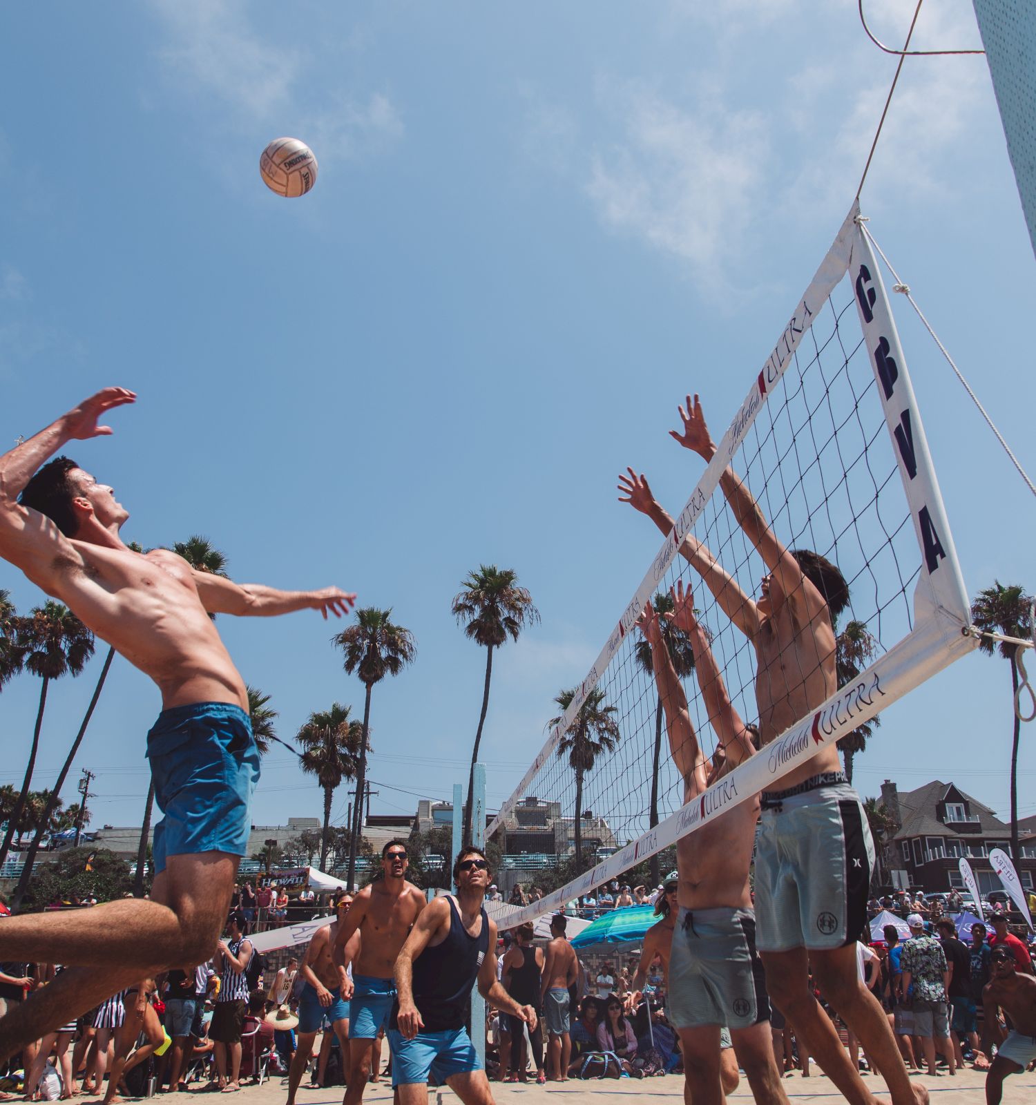 Several people play beach volleyball under a clear sky, with one player leaping to spike the ball as the opposing team attempts to block it.