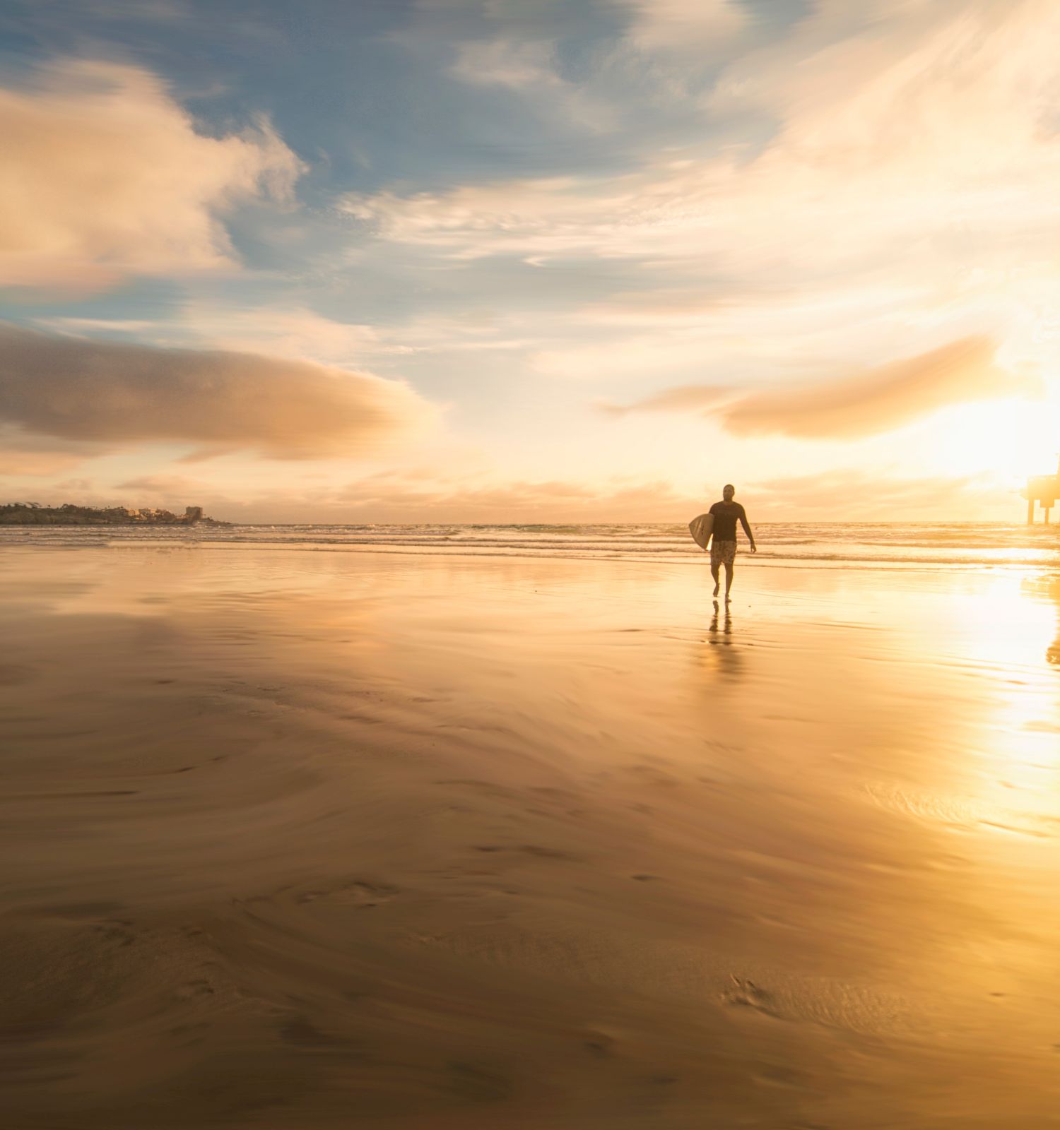 A person is walking on the beach near a pier at sunset, with the sky filled with clouds and the sunlight reflecting off the wet sand.