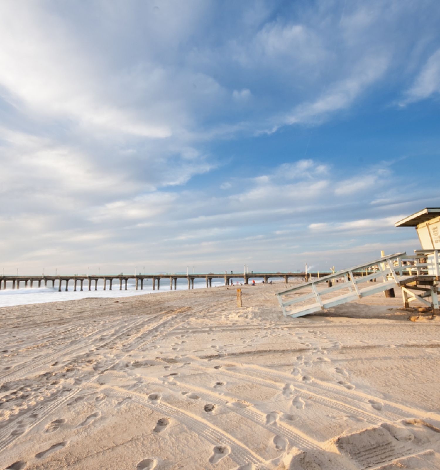 A sandy beach with a lifeguard tower, footprints in the sand, and a long pier extending into the ocean under a partly cloudy sky.