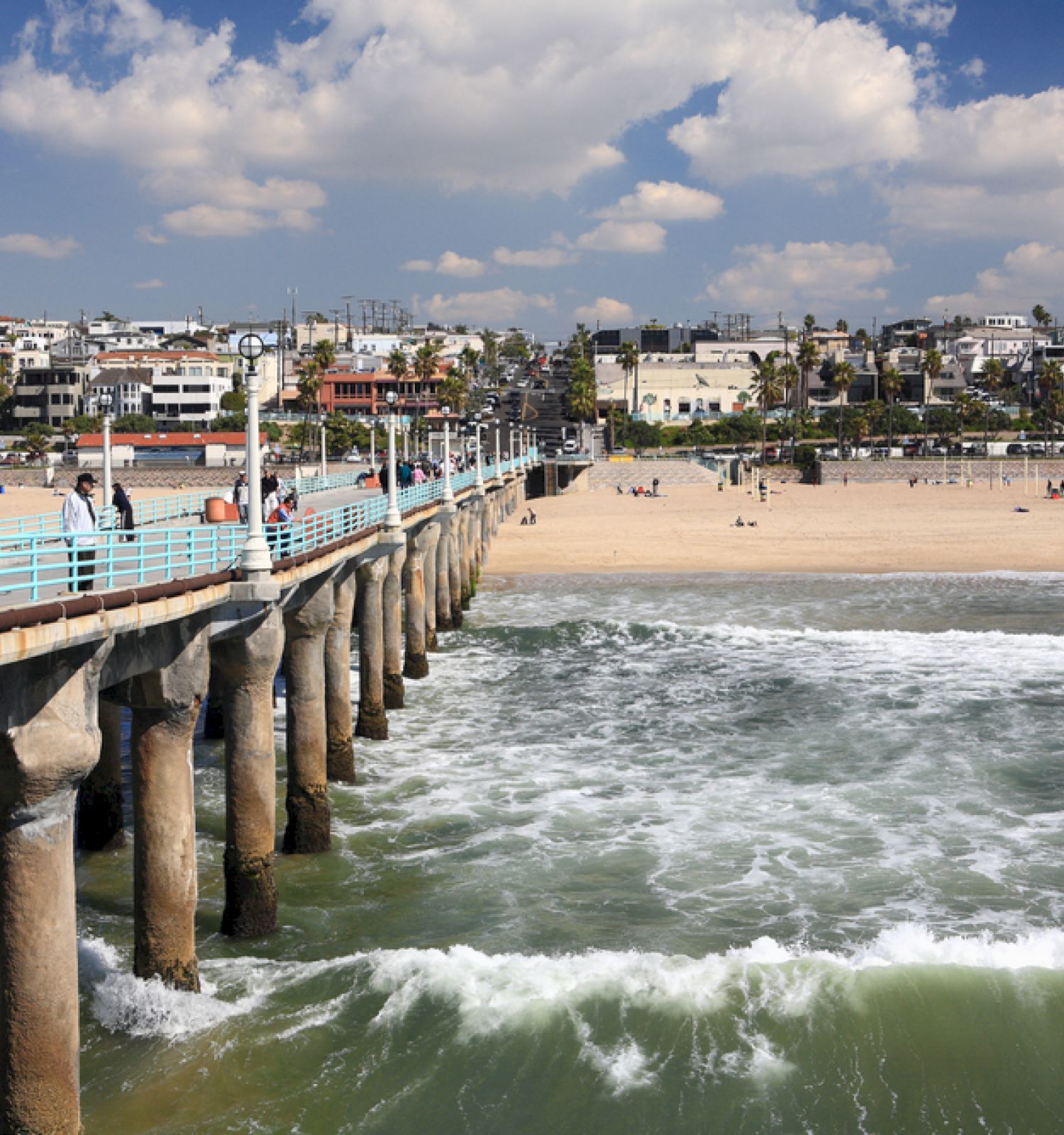 A long pier extends over the ocean with waves crashing below, leading to a sandy beach lined with houses and a partly cloudy sky above it.