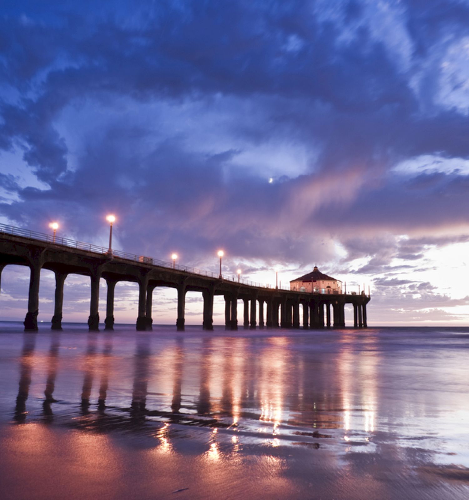 The image shows a pier extending into a calm ocean with a building at the end, under a dramatic, cloudy sky during twilight.