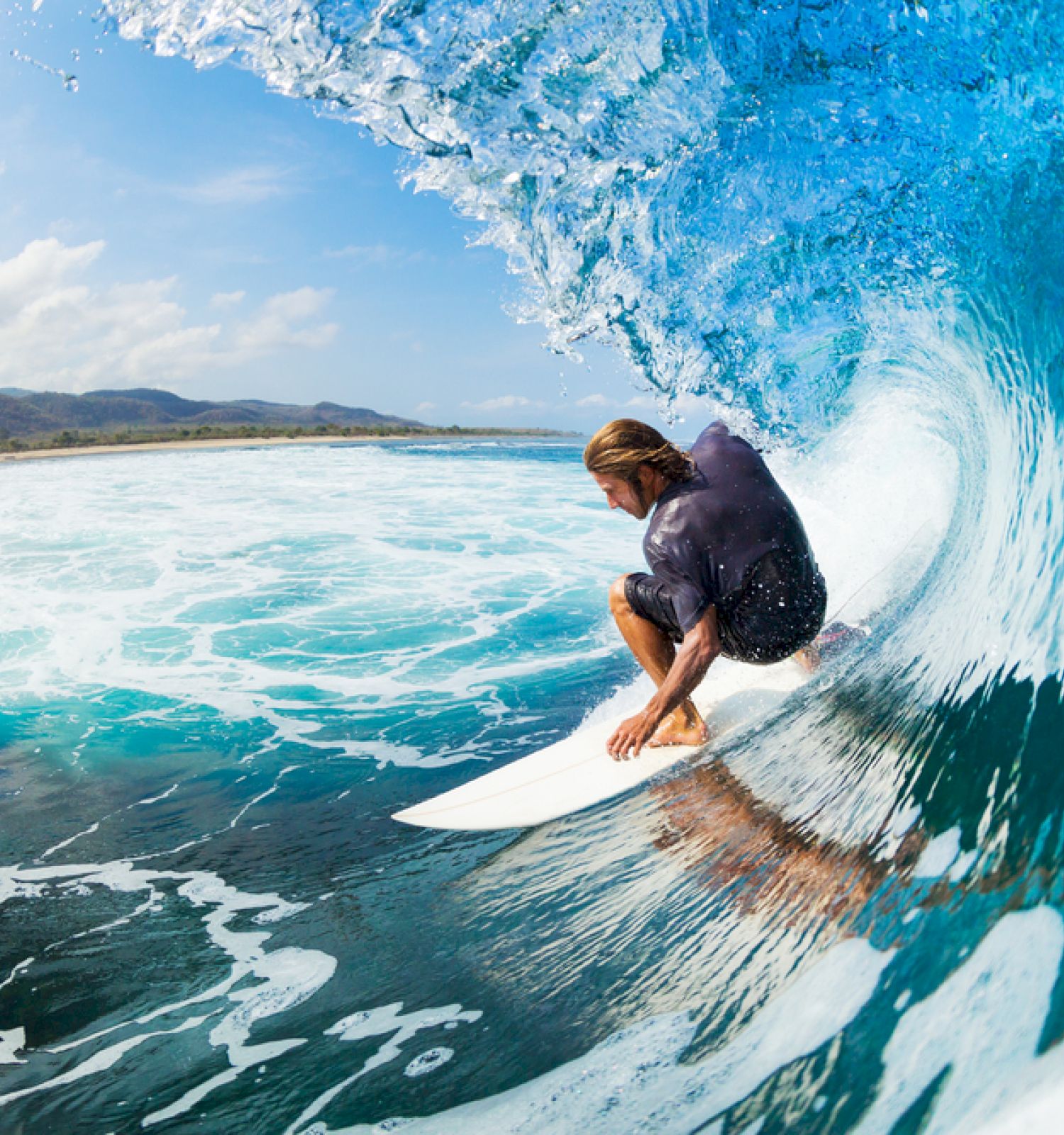 A surfer rides inside a large, blue wave near the shore, with the coastline and mountains visible in the background.