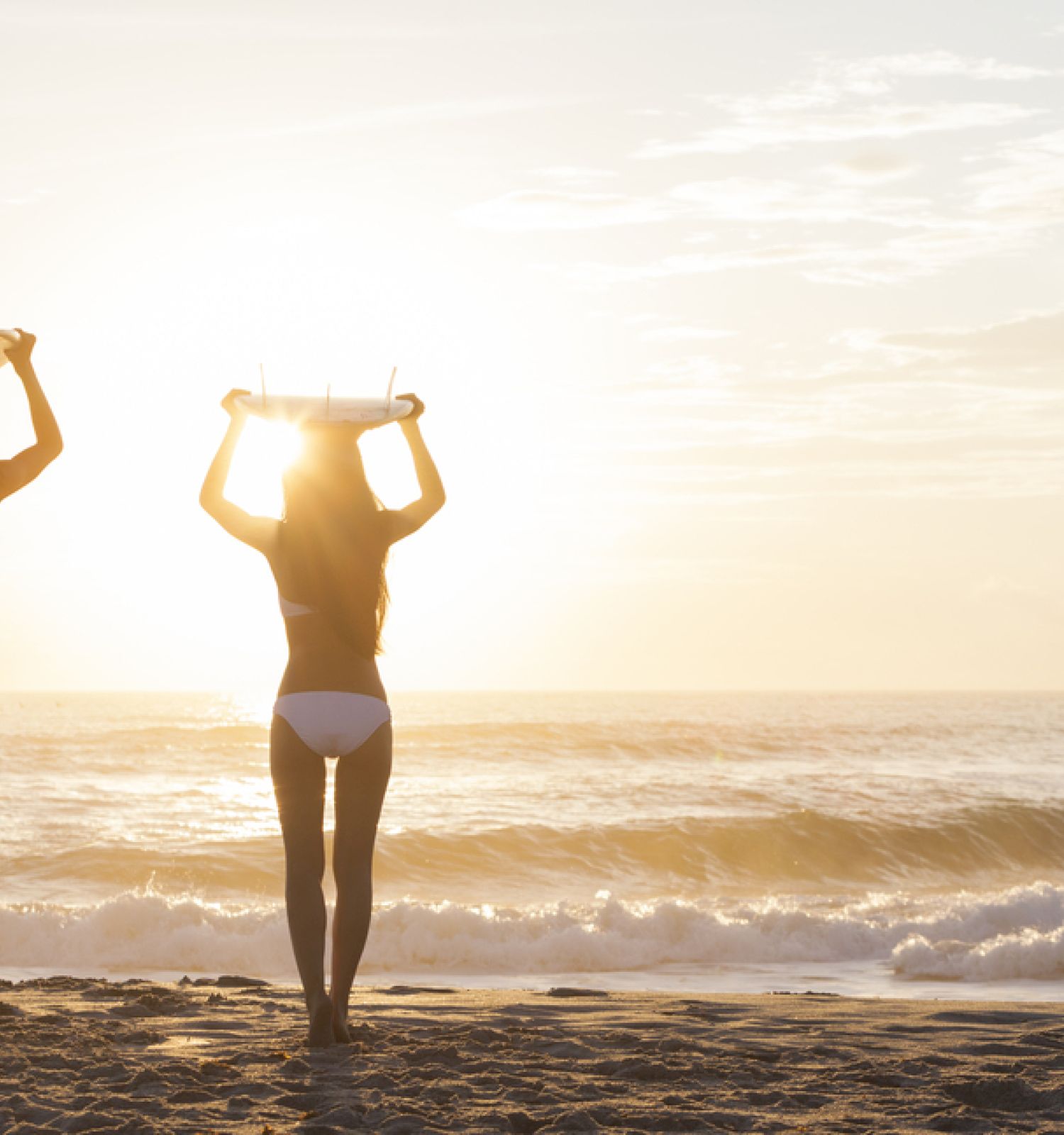 Two people stand on a beach at sunset, holding surfboards above their heads, facing the waves and the horizon.