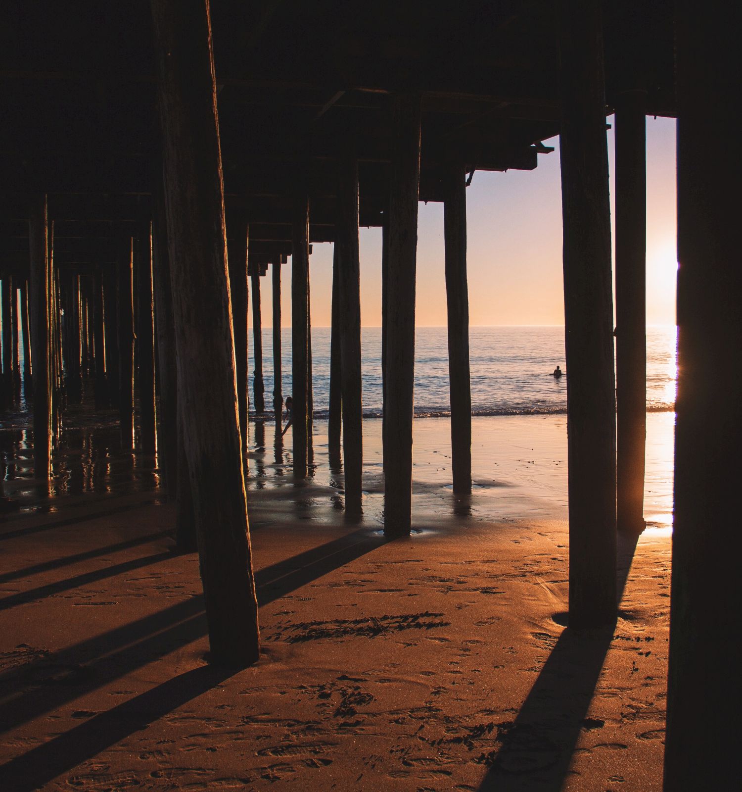 The image shows a view of the beach, looking through the wooden pillars of a pier at sunset, casting long shadows on the sandy shore.