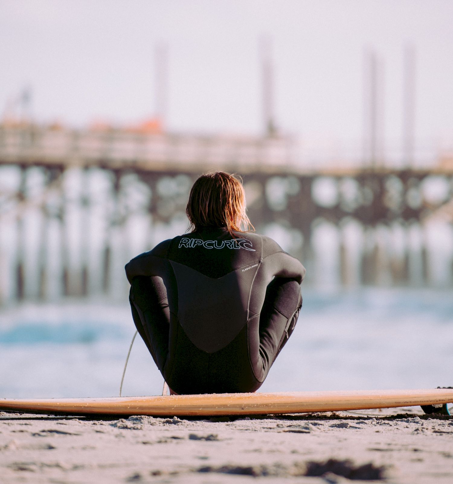 A person in a wetsuit sits on the beach in front of a surfboard, facing the ocean with a wooden pier in the background, at sunrise or sunset.