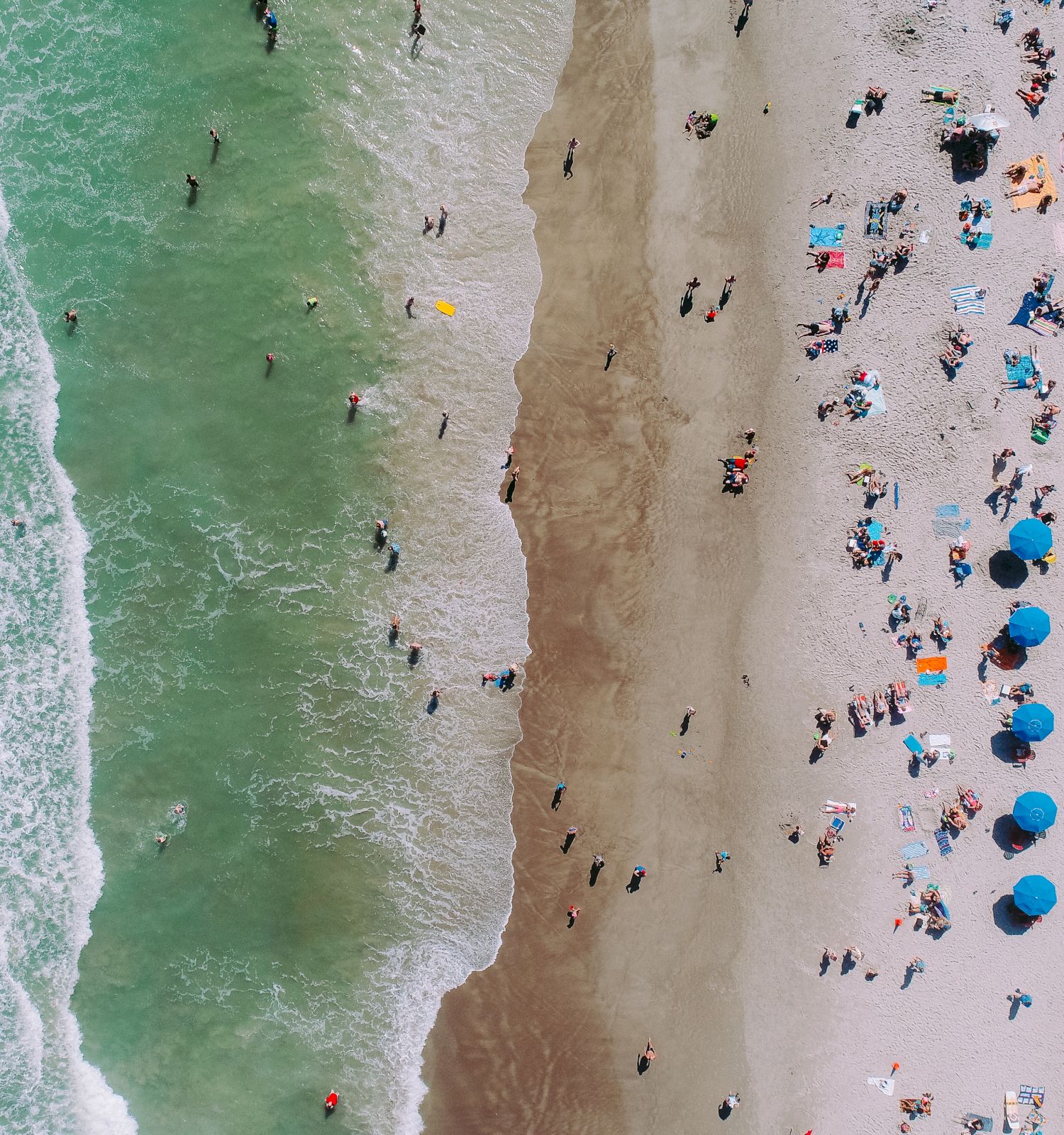 An aerial view of a beach with people swimming in the ocean, and many umbrellas and beachgoers scattered along the sandy shore ending the sentence.