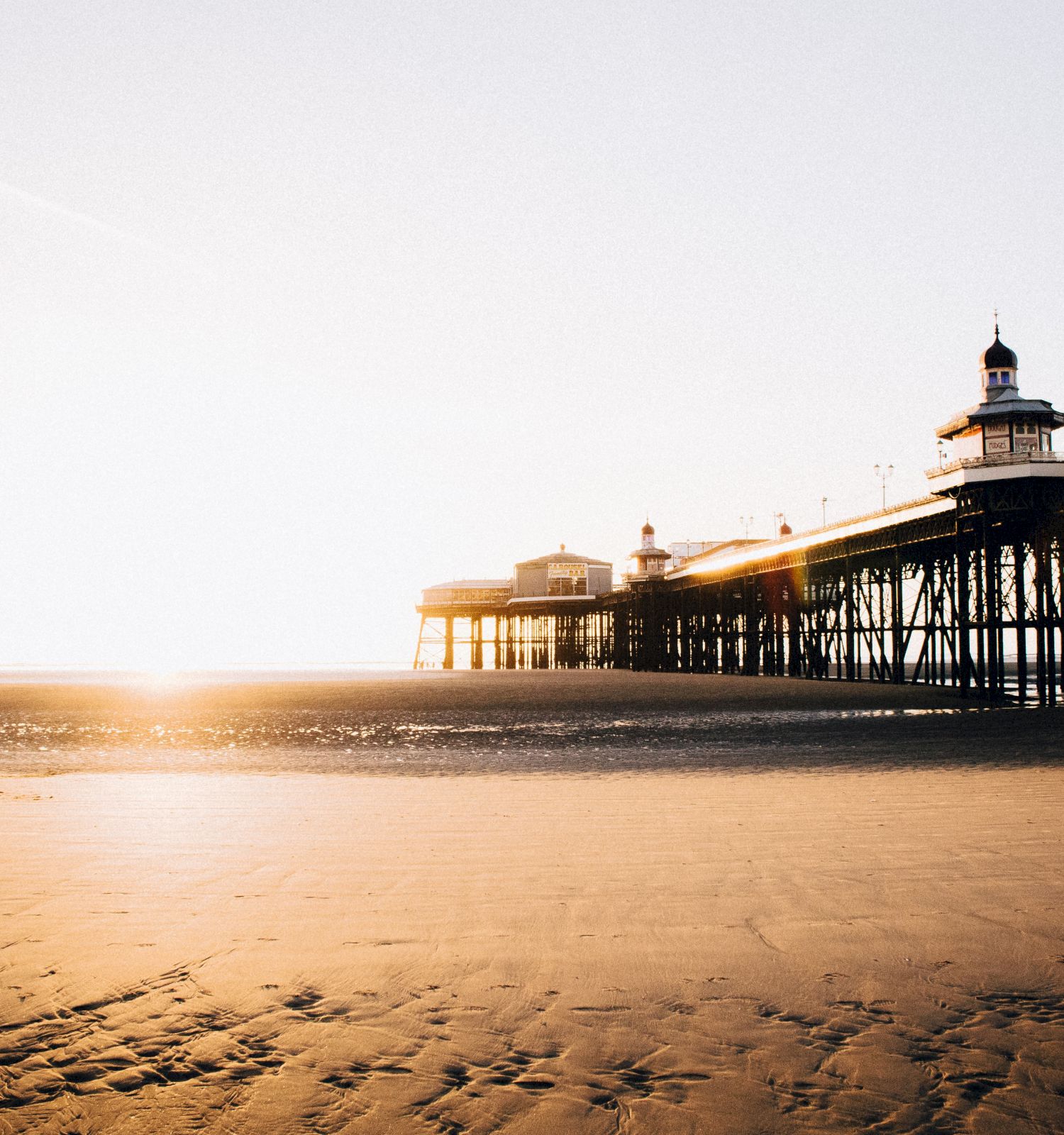 A pier extends over a sandy beach with the sun setting or rising in the background, casting a warm glow on the scene.