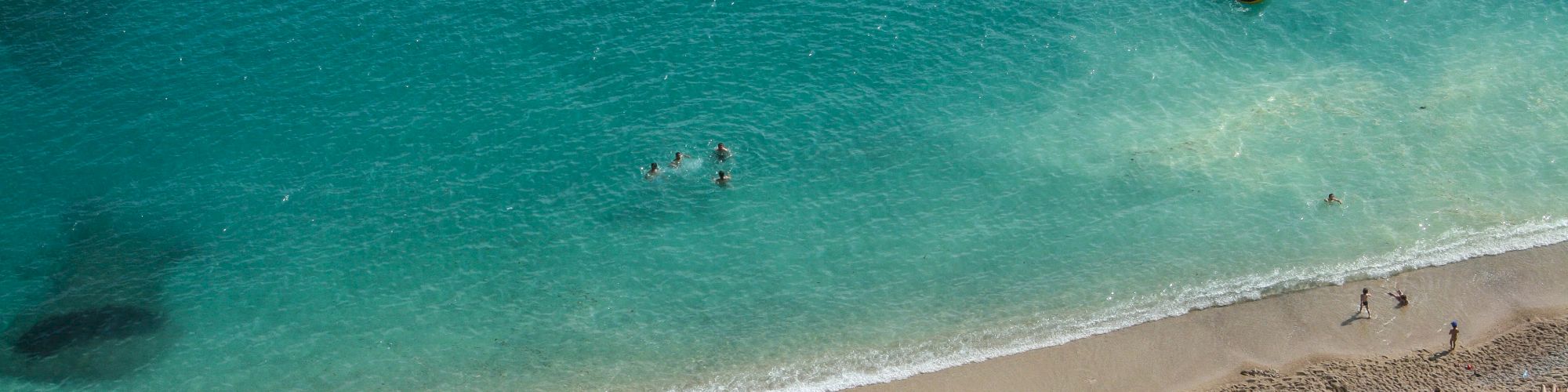 The image shows a sandy beach with people sunbathing and swimming in clear blue water, with a few individuals farther out in the ocean.