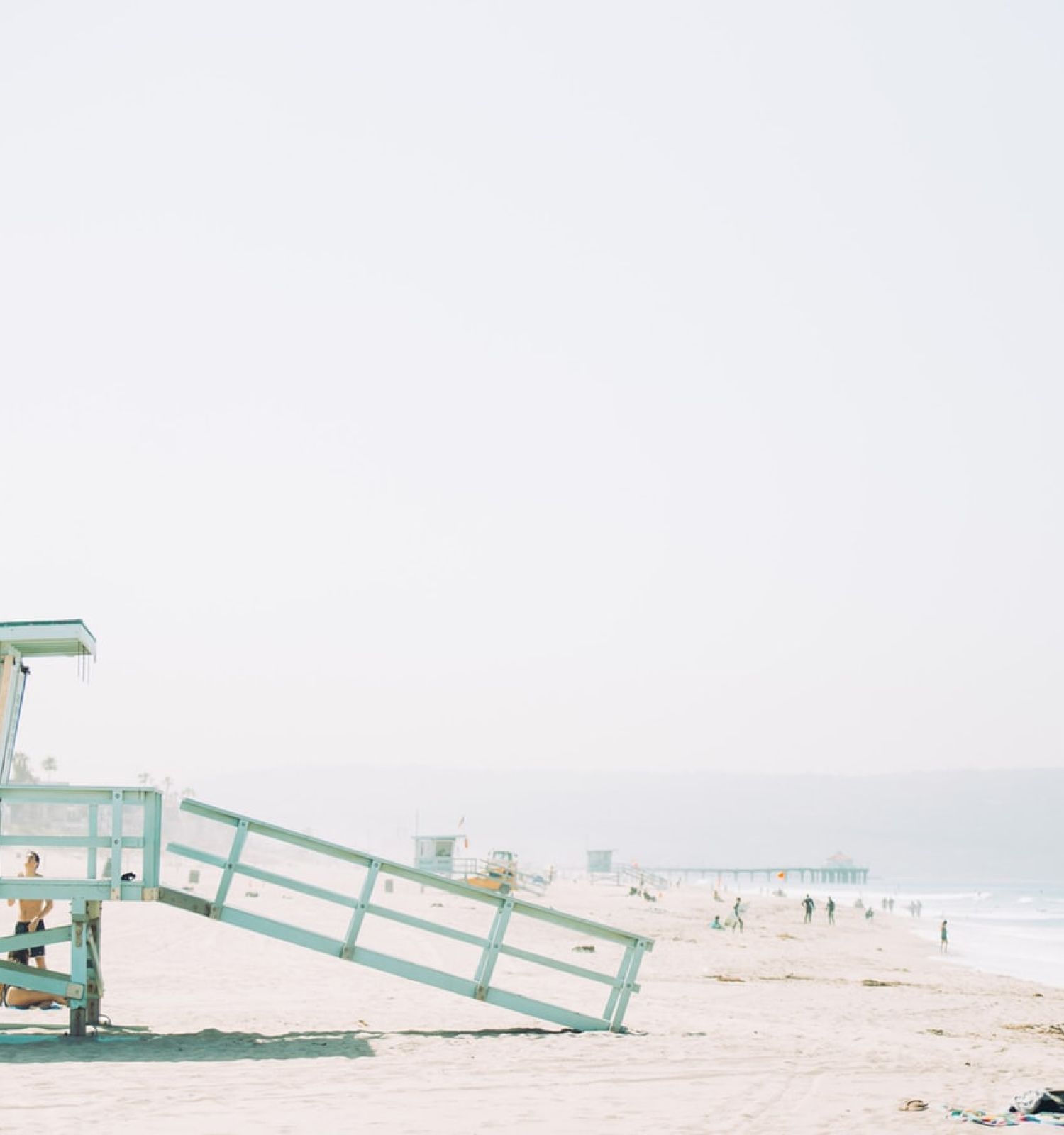 A lifeguard tower is situated on a sandy beach with people in the distance, and a person bending over near the water.