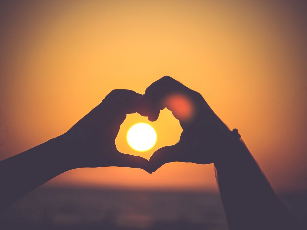 Two hands forming a heart shape frame the setting sun against an orange sky, creating a silhouette effect.