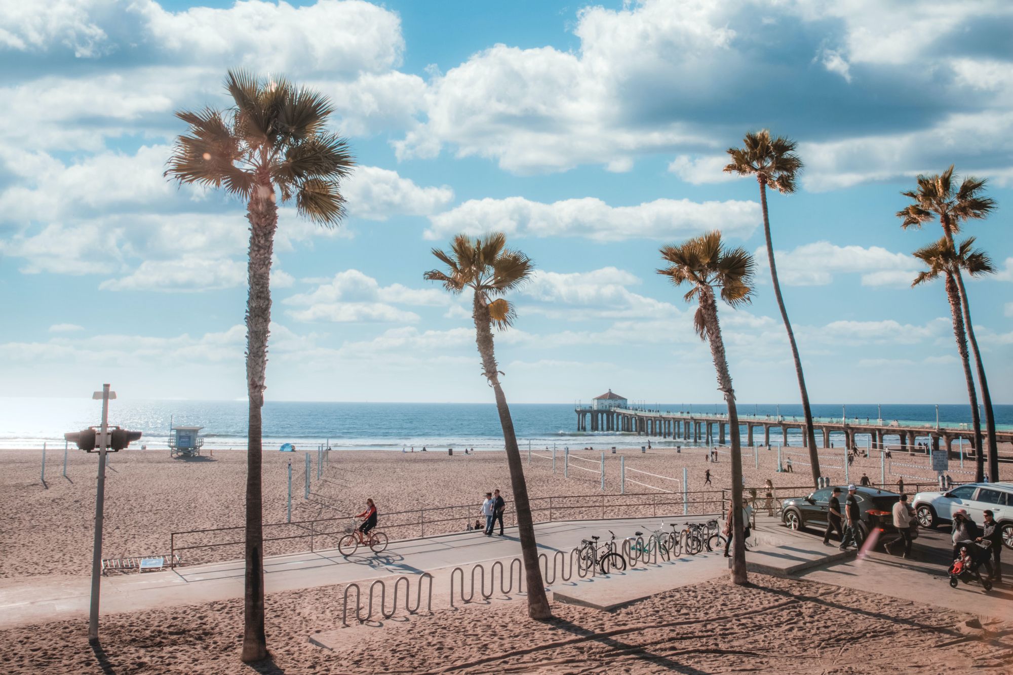 A sunny beach scene with palm trees, a pier extending into the ocean, people biking and walking, and a bike rack in the foreground ends the sentence.