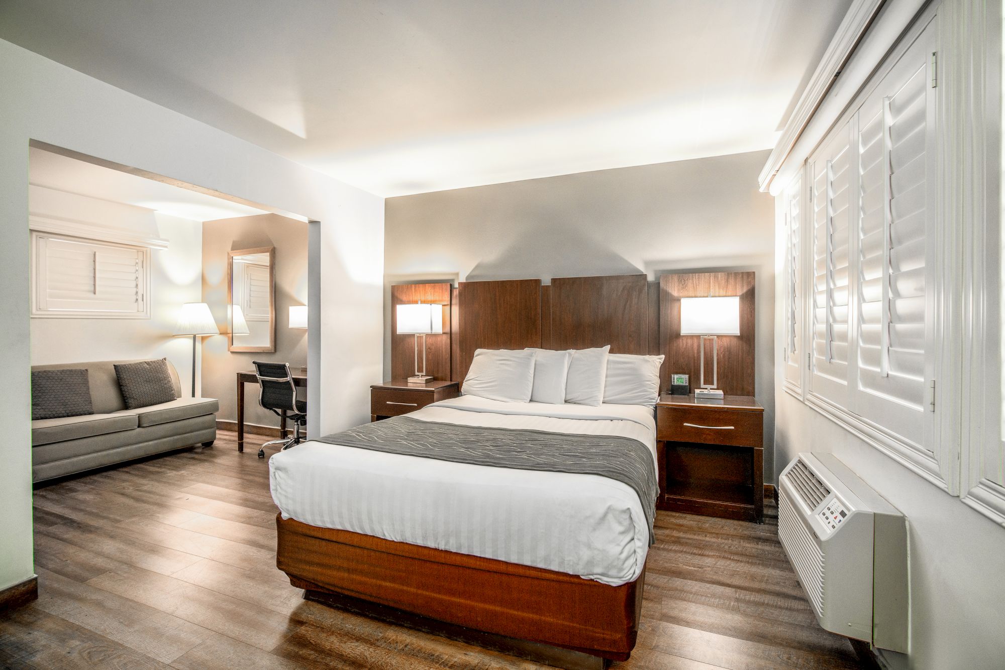 A modern hotel room with a bed, nightstands, lamps, a sofa, a desk area, wooden floors, and white walls.