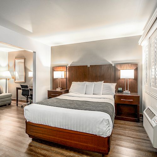 A neatly arranged hotel room with a bed, two bedside tables with lamps, an air conditioner, and a sofa area with a desk and chair in the background.