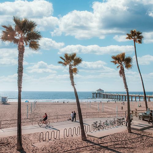 A beach scene with palm trees, a pier, people biking, and blue skies with scattered clouds.