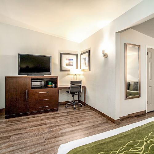 The image shows a hotel room with a TV on a wooden dresser, a desk with a chair and lamp, a tall mirror, an iron and an ironing board.