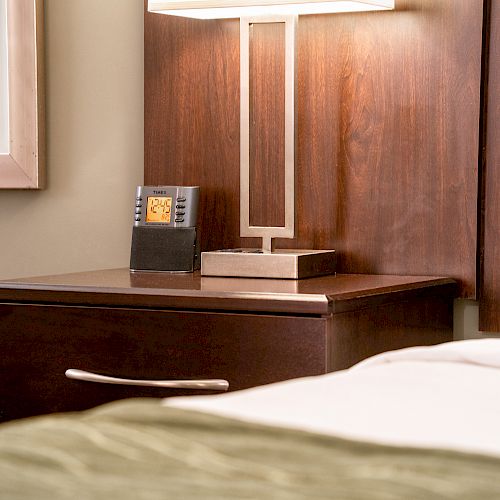 A neatly arranged hotel room with a wooden nightstand, clock, lamp, and a portion of a bed with white linens is shown in this image.