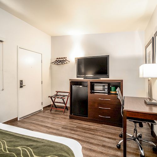 This image shows a hotel room with a bed, TV, microwave, mini-fridge, desk, lamp, and coat hangers on the wall, featuring a clean modern design.