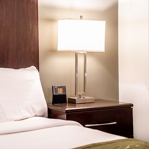 A neatly made bed with white pillows and sheets, a wooden nightstand with a lamp, and a clock, next to a window with shutters.