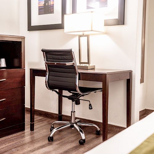 The image shows a minimalist office setup in a room, featuring a sleek black office chair, a wooden desk with a lamp, and framed art on the wall.