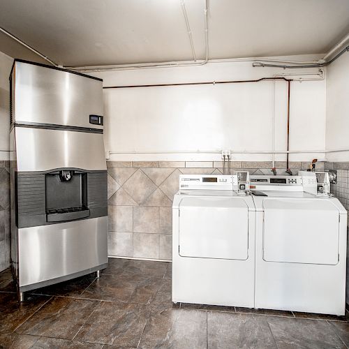 A laundry room with a stainless steel refrigerator, a washing machine, and a dryer against a tiled wall and floor, appears clean and organized.